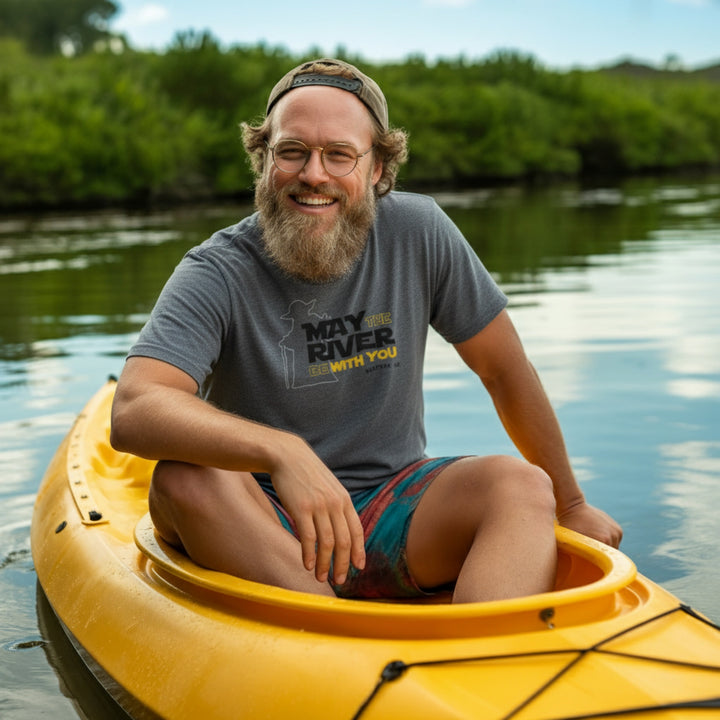 Man with a beard and glasses sitting in a yellow kayak on a calm body of water with greenery in the background. The shirt has the saying May the River be with you referring to the May River in Lowcountry
